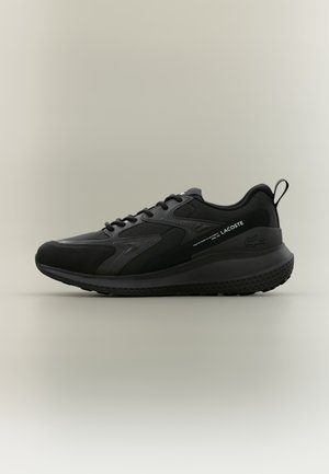 Sort Lacoste sneaker med snørelukning, polstret sål og trækflig på hælen mod en ensfarvet neutral baggrund.