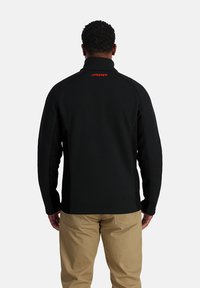 Giacca di pile nera con collo alto, caratterizzata da un motivo testurizzato e il logo rosso "SPYDER" sulla schiena. Abbinata a pantaloni khaki.