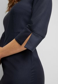 Robe bleu foncé avec un motif à carreaux, dotée de manches trois quarts avec des fentes. Fabriquée dans un tissu texturé et tissé.