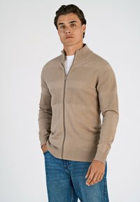 Beżowy sweter zapinany na zamek z teksturowanymi poziomymi pasami, z pr�ążkowanymi mankietami i dołem, wykonany z miękkiego materiału dzianinowego, w zestawieniu z niebieskimi dżinsami.