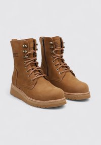 Botas de tobillo de ante marrón con caña alta, cordones en el frente, ojetes metálicos y cremallera lateral. Presentan una suela texturizada y costuras reforzadas.