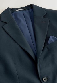 Blazer bleu marine foncé avec revers cranté, bouton unique et pochette assortie dans la poche poitrine sur fond clair.