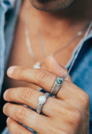 Three rings on a hand: one with a pearl, one textured silver with a green stone, and a detailed silver band, all featuring intricate designs.