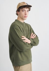 Olivno zelena majica s okroglim izrezom, z rebrastimi manšetami in spodnjim robom, nošena z bež kapo z logotipom. Model stoji s prekrižanimi rokami na enobarvnem ozadju.