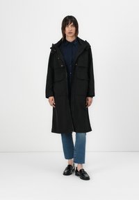 Casaco parka preto com capuz, apresentando dois bolsos frontais, fecho de zip e detalhes em botão, usado sobre uma camisa azul escura de botão e jeans.