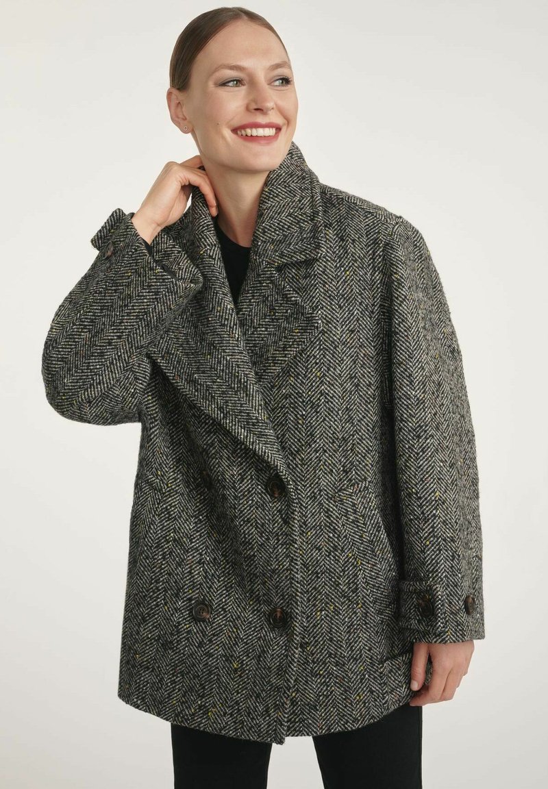 Uśmiechnięta kobieta w szarym płaszczu w jodełkę zapinanym na podwójne guziki z szerokimi klapami, poprawiająca kołnierz jedną ręką, stojąca na jednolitym tle.