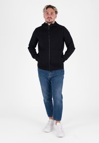 Felpa nera con zip e collo alto, abbinata a jeans di jeans azzurri e sneakers bianche. Tessuto morbido, silhouette aderente e design funzionale.