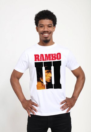 Man glimlacht met handen op de heupen, draagt een wit T-shirt met rode "RAMBO" tekst en groot zwart Romeins cijfer III met gezicht erin.