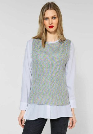 Grå texturerad stickad ärmlös topp lagd över en ljusblå långärmad blus, kombinerad med svarta jeans.