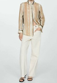 Persona che indossa una camicia a righe beige e bianche con bottoni, pantaloni bianchi a vita alta e gamba ampia, e tacchi aperti neri, con una mano in tasca.