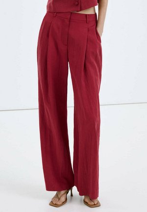 Pantalones rojos de talle alto y pierna ancha con pliegues delanteros, combinados con sandalias de tacón marrón claro, mostrados desde la cintura hasta los pies con un fondo liso.