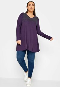 Lila långärmad tunika med en strukturerad, prydnadssmyckad halsringning, som är kombinerad med mörka slim fit jeans och vita sneakers.