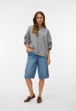 Donna che indossa una felpa grigia oversize, pantaloncini in denim blu fino al ginocchio e scarpe marroni con lacci, in piedi con una mano in tasca.