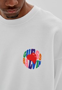 Witte sweatshirt met een kleurrijk rond logo met de tekst "GUESS BALVIN" en een rode hart in het midden. Zachte textuur, geribbelde kraag.