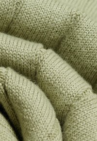 Gros plan d’un tissu tricoté doux vert clair avec texture et fibres visibles, montrant des plis et des ombres douces.
