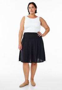 Jupe noire froncée avec une coupe décontractée, associée à un débardeur blanc ajusté. Le modèle porte des ballerines tissées beiges. Texture du tissu lisse.