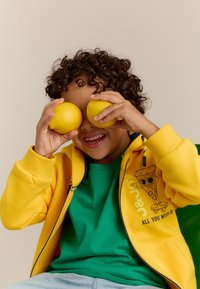 Sweat-shirt zipé jaune avec un graphique de pizza, t-shirt vert en dessous, et cheveux bouclés. Tient deux citrons devant les yeux, souriant.