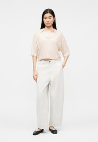 Frau steht und trägt eine lockere, blassrosa Bluse mit kurzen Ärmeln, hoch taillierte, weite weiße Hose und schwarze flache Schuhe.