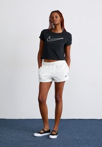 Mulher com tranças vermelhas vestindo um top cropped preto da Nike, calções brancos da Nike e sapatilhas Mary Jane pretas, de pé com as mãos nos bolsos sobre um tapete azul.