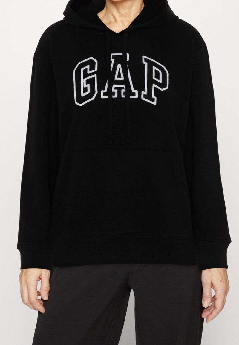 Sudadera negra con capucha que presenta el logo "GAP" bordado en blanco, bolsillo canguro frontal y puños y dobladillo acanalados. Textura de tela suave.
