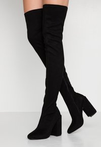 Botas de ante negras por encima de la rodilla con un tacón bloque ancho, textura suave, punta afilada y detalle de cremallera lateral.