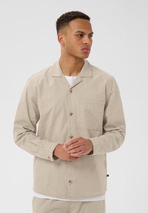 Mies, jolla on lyhyet kiharat hiukset, beige nappipaita ja valkoinen aluspaita, seisoo kädet ristissä ja katsoo oikealle.