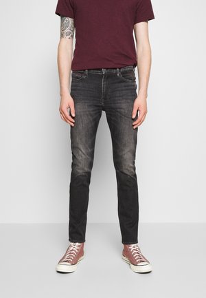 Homme portant un jean slim noir délavé, un t-shirt bordeaux, des baskets montantes roses, debout les bras détendus le long du corps.