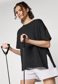 Crna oversized majica kratkih rukava, u kombinaciji s bijelim sportskim kratkim hlačama. Model drži crnu vježbačku opremu s teksturiranim ručkama.