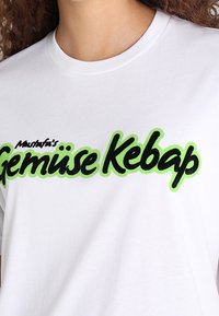 Bílý bavlněný tričko s černým a neonově zeleným textem, který zní "Mustafa's Gemüse Kebap" v tučném, hravém písmu.