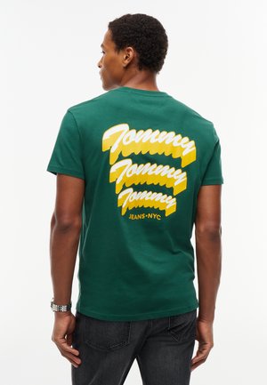 Homme portant un t-shirt vert avec un texte "Tommy Tommy Tommy Jeans NYC" jaune et blanc dans le dos, associé à un jean foncé et une montre argentée.