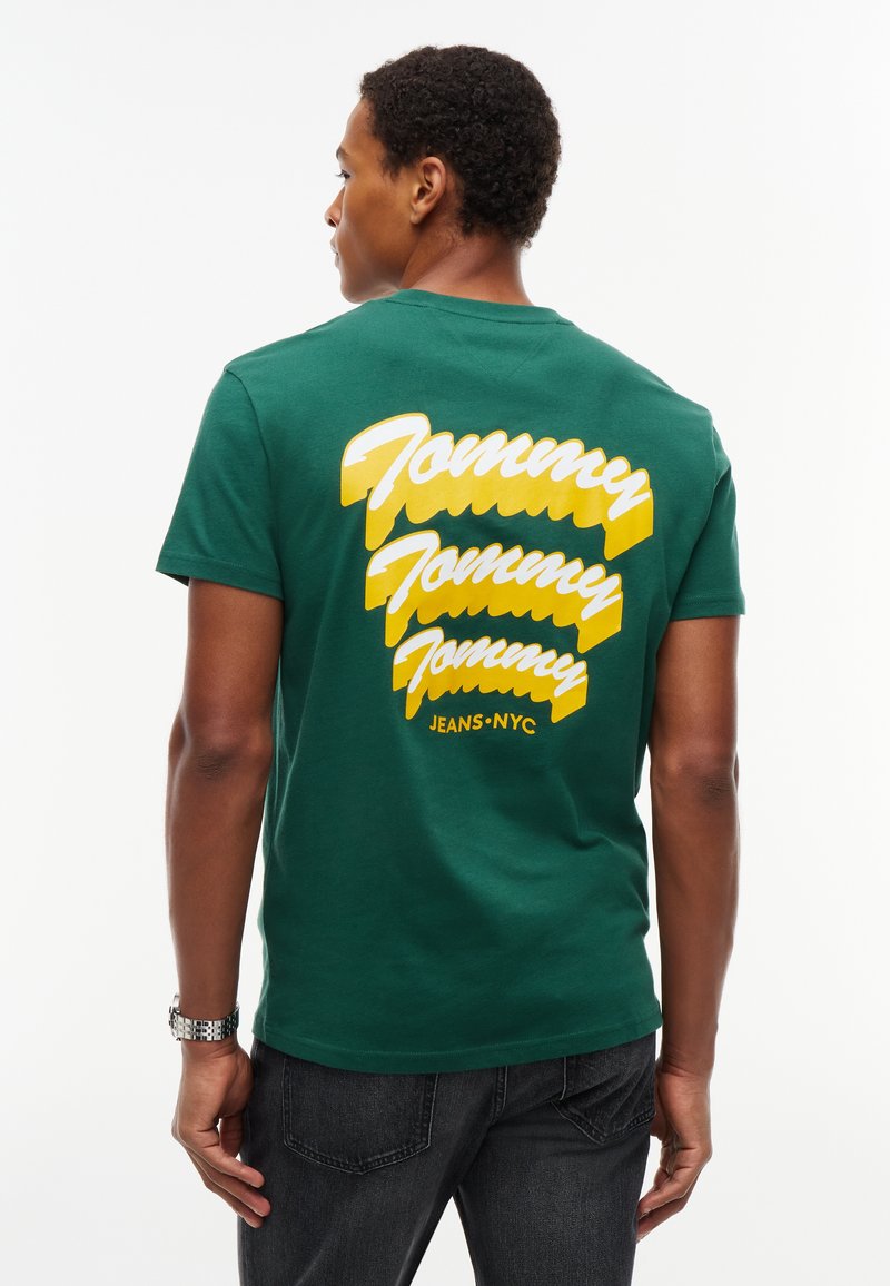 Άνδρας που φοράει πράσινο t-shirt με κίτρινο και λευκό κείμενο "Tommy Tommy Tommy Jeans NYC" στην πλάτη, σε συνδυασμό με σκούρο τζιν και ασημένιο ρολόι.