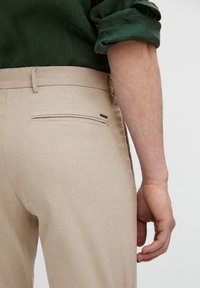 Prawa ręka i dłoń osoby obok beżowych spodni z tylną kieszenią oraz ciemnozielonej koszuli z długim rękawem włożonej w spodnie.