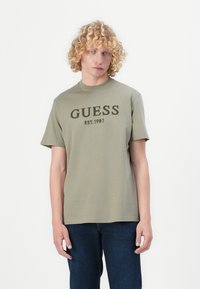 Maslinasto zelena pamučna majica s kratkim rukavima, na kojoj se čitko ističe natpis "GUESS EST.1981" u tamnozelenoj boji preko grudi. Jednostavan dizajn, okrugli izrez.