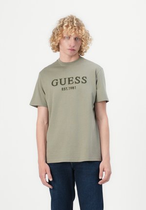 Tricou din bumbac verde măsliniu, cu mâneci scurte, având inscripția "GUESS EST.1981" în verde închis pe piept. Design simplu, cu guler rotund.