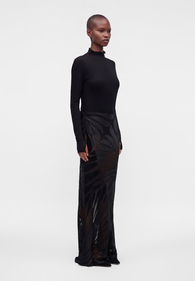 Femme debout de profil, vêtue d’un col roulé noir moulant et d’une longue jupe noire transparente à motifs abstraits, sur fond uni.