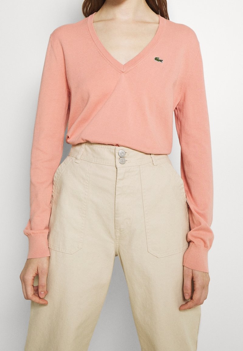 Femme portant un pull rose clair à col en V et manches longues avec un petit logo vert, rentré dans un pantalon beige taille haute à boutons.