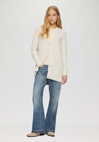 Helle, crèmefarbene Strickjacke mit Knöpfen, offenem Vorderteil und gerippten Bündchen, kombiniert mit blauen Schlagjeans und beigen Schuhen. Glattes, strukturiertes Material.