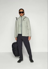 Veste imperméable gris clair avec un col montant et une fermeture éclair devant, superposée à un haut assorti. Un pantalon cargo noir et des bottes robustes complètent la tenue.