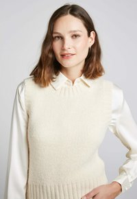 Gilet en tricot beige clair en matériau doux, avec un ourlet côtelé et un col montant, superposé à une blouse blanche à col.