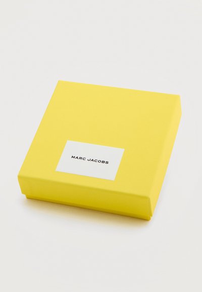 Κίτρινο ορθογώνιο κουτί με λευκή ετικέτα στο κέντρο επάνω, που εμφανίζει το κείμενο «MARC JACOBS.»