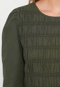 Haut vert avec un design smocké texturé et du tissu froncé, un col rond et des manches longues. Matériau lisse et doux.
