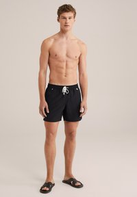 Schwarze Schwimmshorts mit einem Kordelzugbund, aus leichtem Material, mit einem kleinen Logo-Patch und kombiniert mit schwarzen Sandalen.
