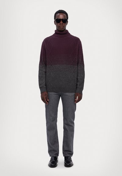 Pull à col roulé en maille dans un dégradé bordeaux avec des motifs texturés, associé à un jean gris et des chaussures noires. Le modèle porte des lunettes de soleil.