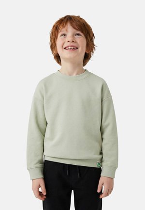 Jeune garçon roux portant un sweat-shirt gris clair et un pantalon noir, souriant et regardant vers le haut sur un fond uni.