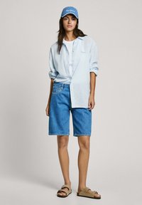 Hellblau gestreifte Button-Up-Bluse, weißes T-Shirt, Denim-Shorts und braune Sandalen mit offenen Zehen. Blauer Cap mit besticktem Text.