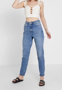 Valkoinen lyhyt toppi, jossa olkaimet ja metallikoristeet, yhdistetty vaaleansinisiin korkeavyötäröisiin farkkuihin ja mustiin sandaaleihin, joissa on pyöreitä niittejä.