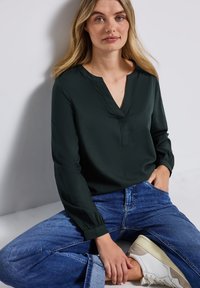 Mörkgrön blus med lång ärm och V-ringning, slät textur och avslappnad passform, i kombination med blå jeans och vita sneakers.