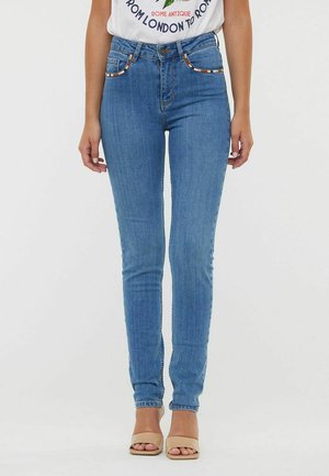 Jean slim bleu taille haute, avec des coutures contrastées sur les poches et une texture subtilement décolorée. Associé à des sandales à talons beiges.