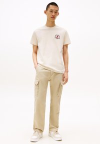 Beige t-shirt with logo on chest, paired with light beige cargo pants featuring side pockets. Model wears white sneakers.