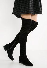 Černé nadkolenky vyrobené z měkkého semiše, s plochým podpatkem, bočním zipem a hladkou texturou. V doprovodu kostkované sukně.
